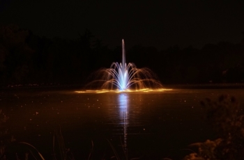 The 2.0hp Aqua Control Fleur De Lis floating fountain is highlighted by blue LED lights and situated in an evening setting