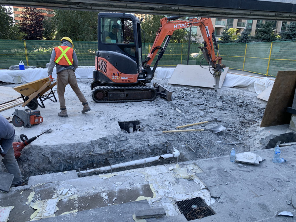 Small excavator with a demolition hammer removing the fountain, with concrete almost thirty inches deep